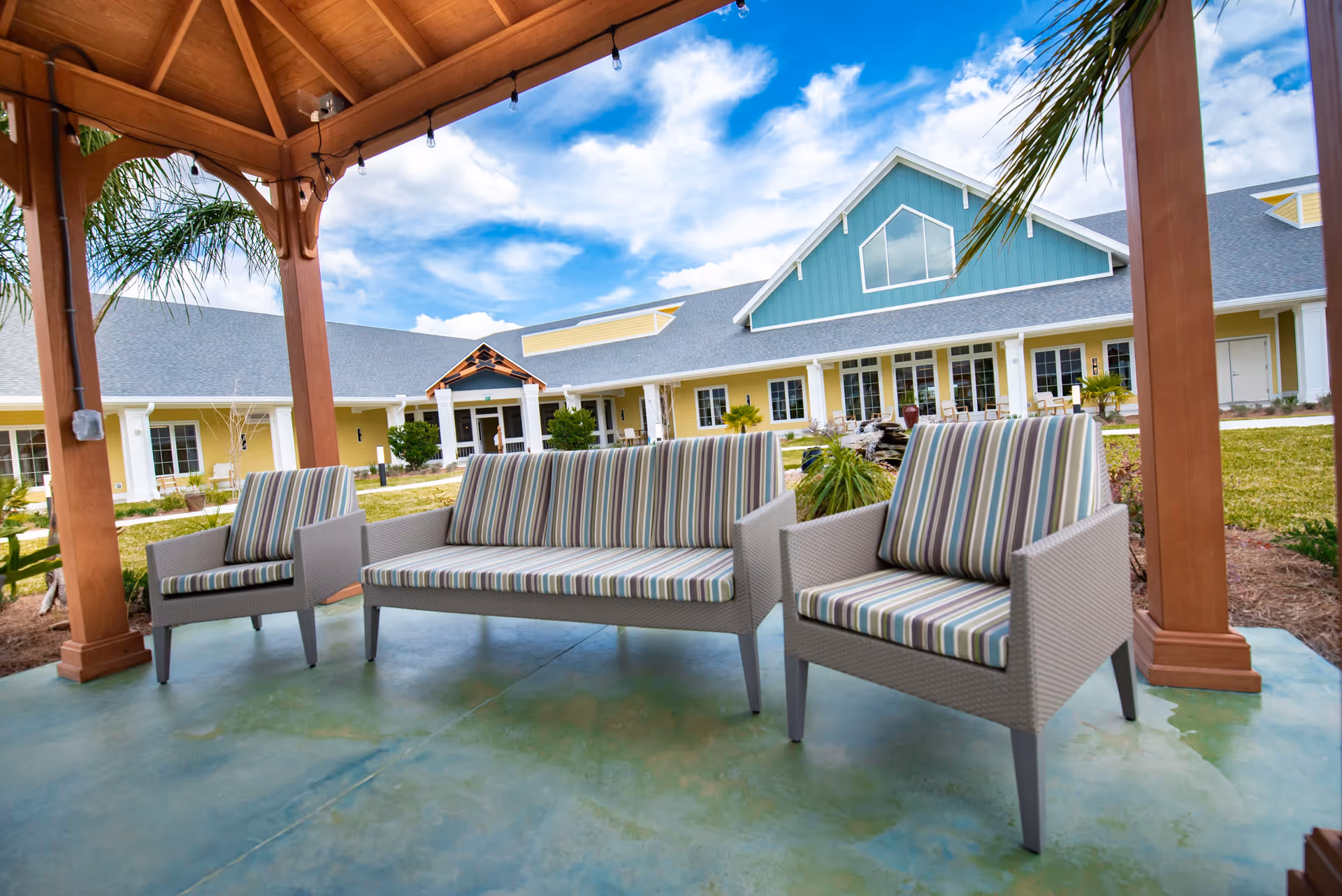 Striped-cushioned outdoor sofa and two chairs under a wooden pavilion with the facility building in the background.