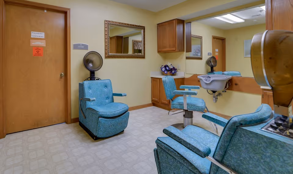 Interior view of a senior living facility's hair salon area with vintage blue salon chairs, a hair dryer, a wash basin, a large mirror on the wall, wooden cabinets, and a closed wooden door with signs on it.