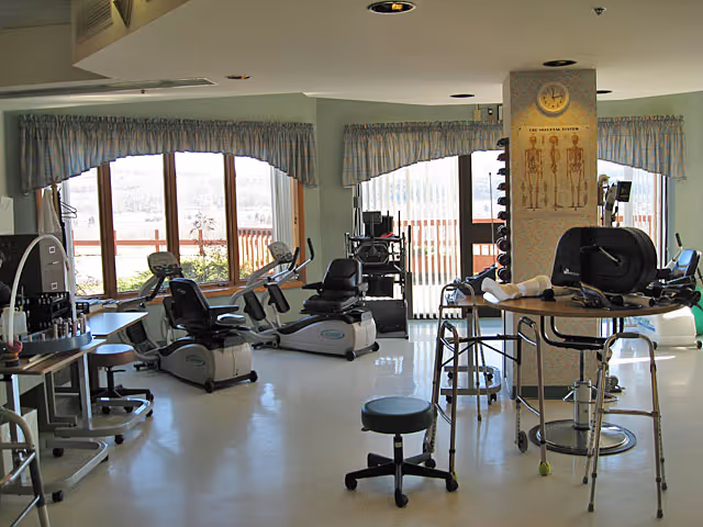 Rehabilitation gym room with recumbent exercise bikes, therapy equipment, weights, stools and large windows.