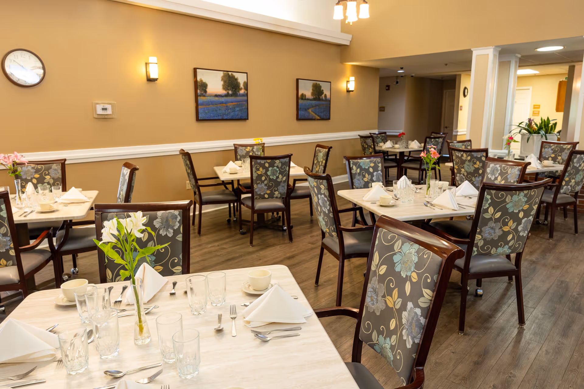 A dining room in a senior living facility with several tables set with white napkins, cups, glasses, and silverware. Each table has a small vase with flowers. The chairs have floral patterned upholstery. The walls are beige with two landscape paintings and wall sconces providing light. A clock is visible on the wall.