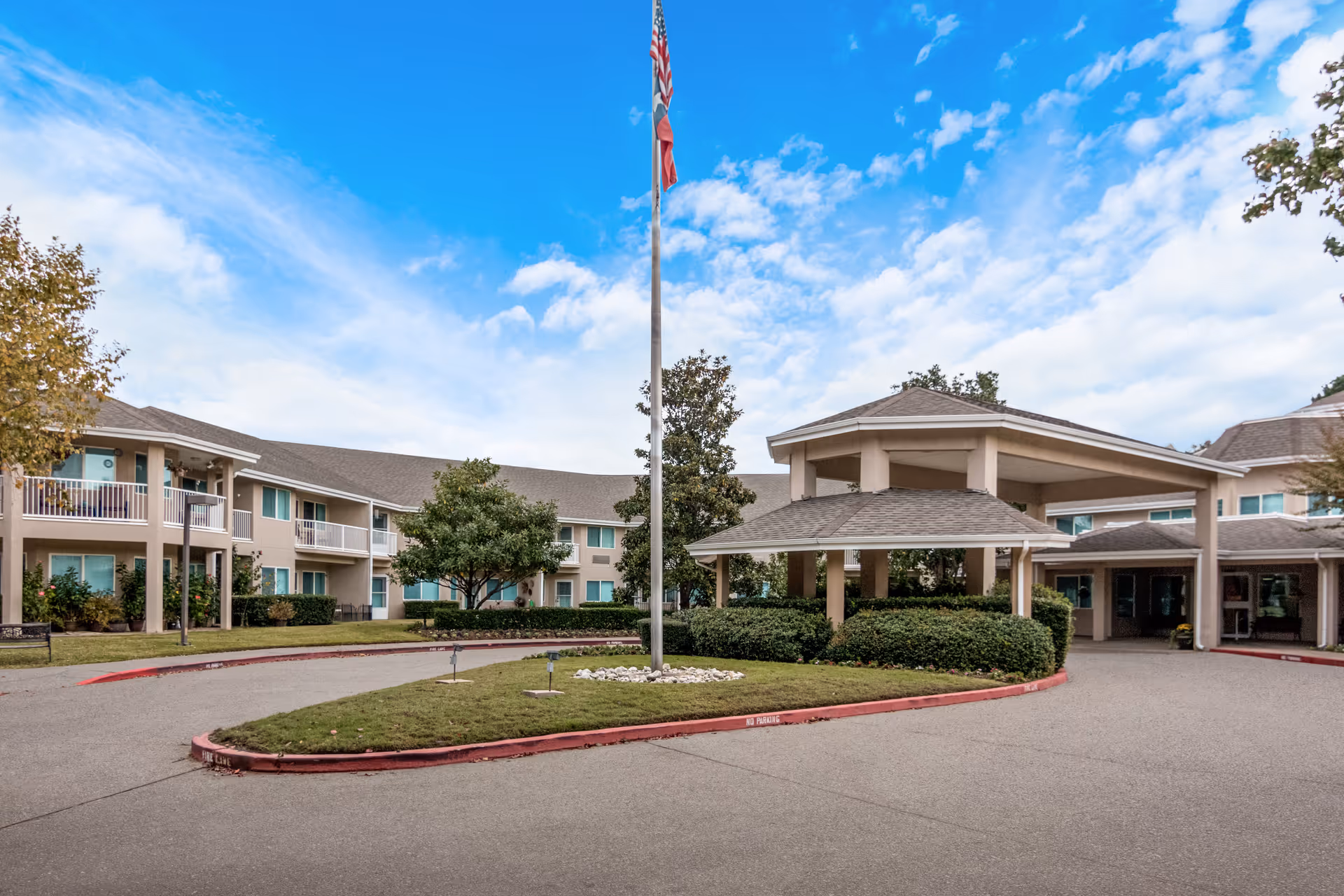 Exterior view of Asher Point Independent Living of Tyler showing a two-story building with balconies, a covered entrance with columns, a flagpole with an American flag, and landscaped greenery under a partly cloudy blue sky.