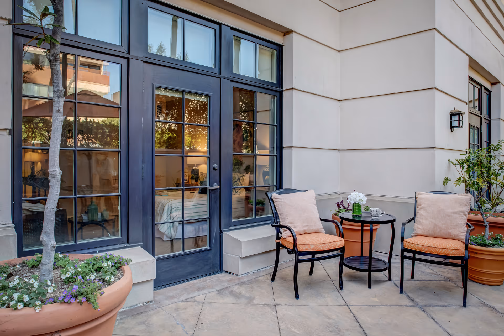 Outdoor patio area with two cushioned chairs and a small round table between them, placed on a tiled floor. The patio is adjacent to a building with large glass doors and windows, through which a bedroom with a bed and lamp is visible. There are large potted plants on either side of the seating area.