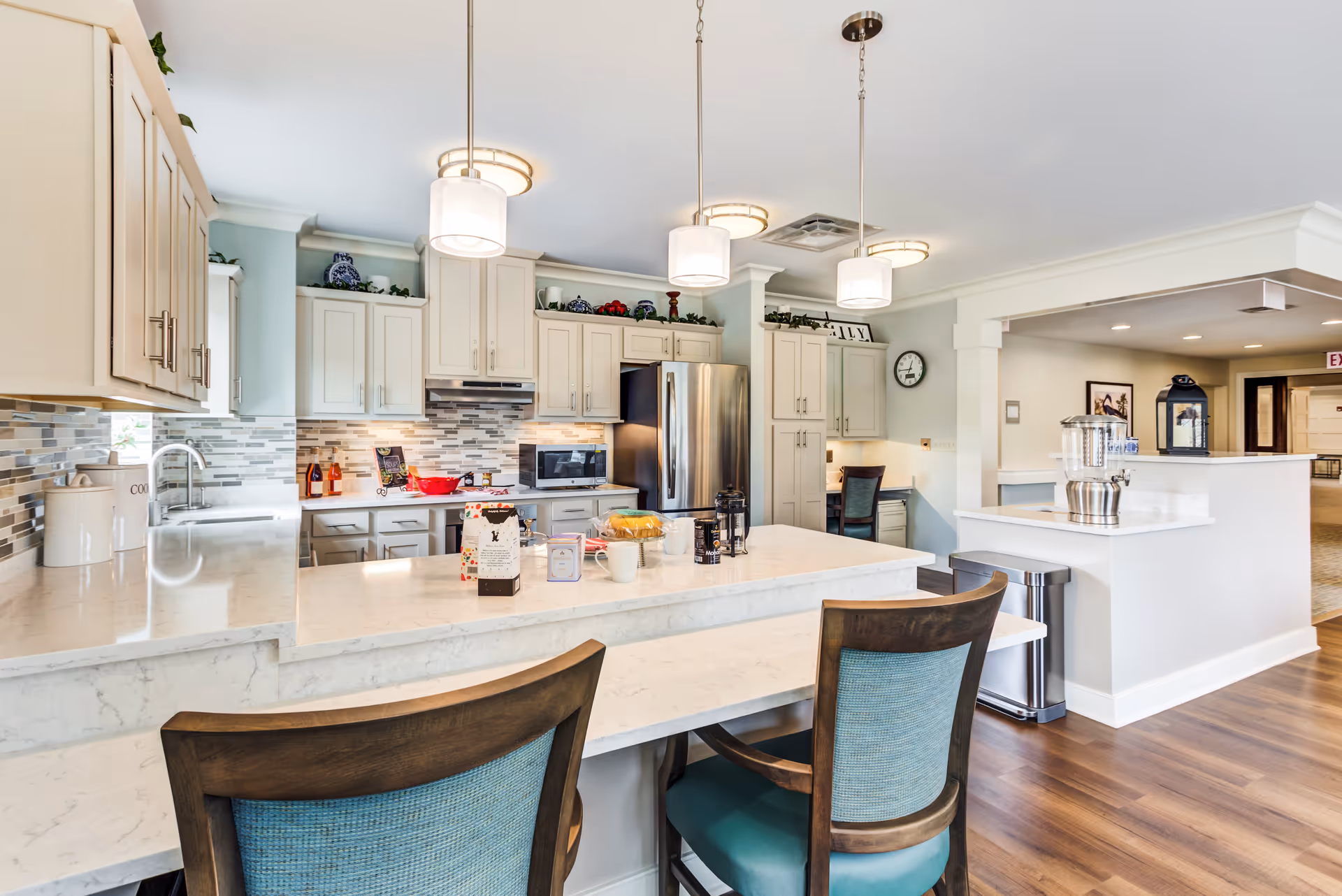 Bright and modern kitchen area with white cabinetry, stainless steel refrigerator, microwave, and a tiled backsplash. A marble countertop island with two wooden chairs featuring blue upholstery is in the foreground. Pendant lights hang from the ceiling, and there is a small desk area with a chair in the background. The floor is wooden, and the space opens into a hallway.