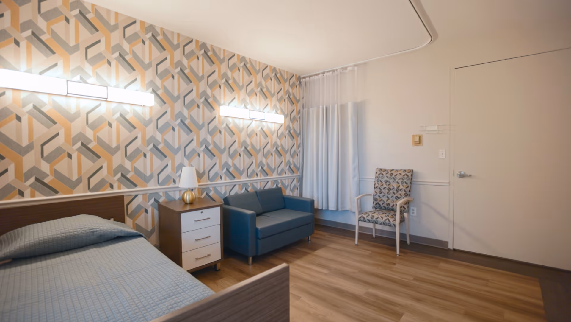A senior living facility room with a single bed covered in a blue quilt, a wooden nightstand with a lamp, a small blue loveseat, and a patterned armchair. The wall behind the bed and loveseat has a geometric patterned wallpaper, and the floor is wood. There are two horizontal lights on the patterned wall and a white curtain covering a window.