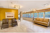 A spacious and well-lit common area with large windows along one wall letting in natural light. The room features a mustard yellow accent wall with a framed picture, two brown leather chairs, a beige sofa, and two modern ceiling light fixtures. The floor is light-colored and the overall decor is clean and minimalistic.