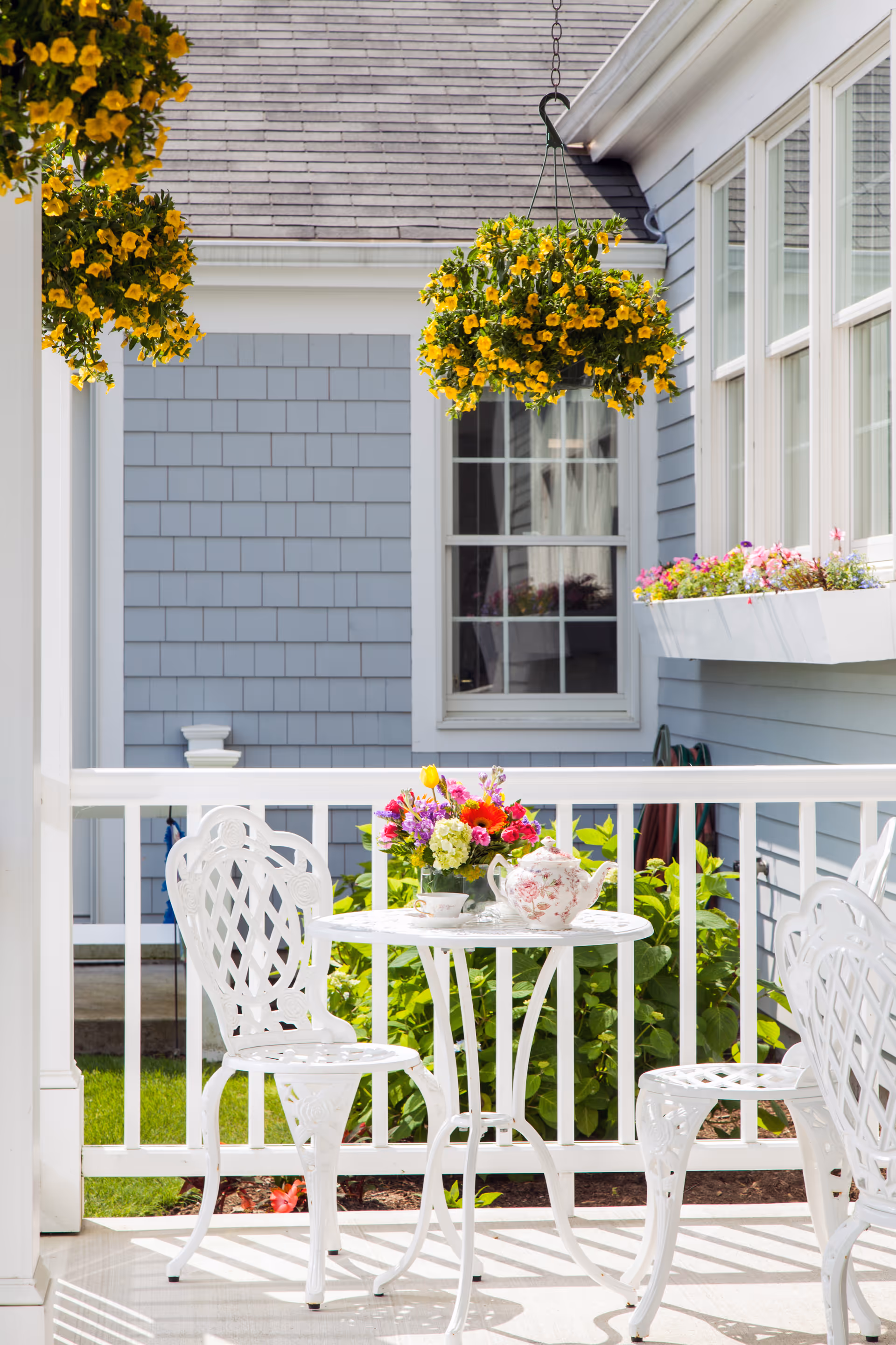 A bright outdoor patio area with white metal chairs and a small round table. The table holds a floral teapot, a teacup, and a colorful bouquet of flowers. Yellow hanging flower baskets and a window box with pink flowers adorn the light blue exterior wall of the building.