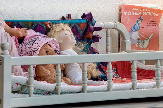 A small white wooden crib or bed holding a blonde-haired doll, a teddy bear wearing a pink bonnet, and a colorful blanket. Behind the crib, there is a book titled 'Baby's First Mother Goose' with an illustration of a witch riding a goose.