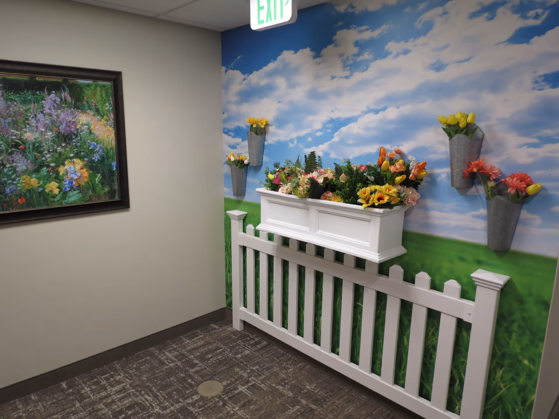 Indoor corner decorated with a white picket fence and a white flower box filled with colorful artificial flowers. The wall behind features a mural of a blue sky with clouds and green grass. There are also metal wall-mounted flower holders with artificial flowers. A framed painting of a garden scene hangs on the adjacent plain wall. An exit sign is visible above.
