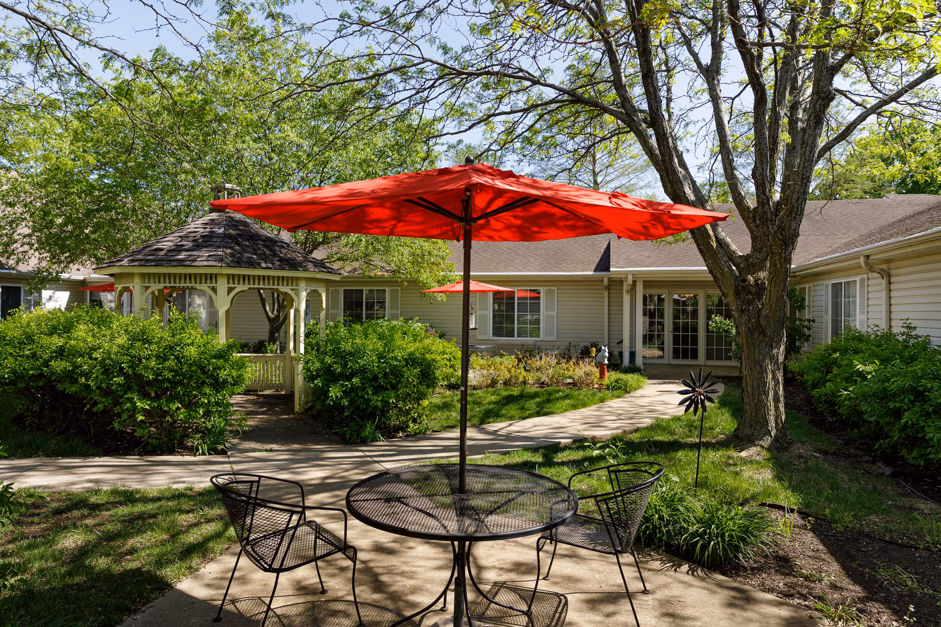 Outdoor patio area at Homestead of Olathe Memory & Dementia Care featuring a round metal table with four matching chairs under a large red umbrella. In the background, there is a white gazebo surrounded by green bushes and trees, with a beige building and windows visible behind the greenery.