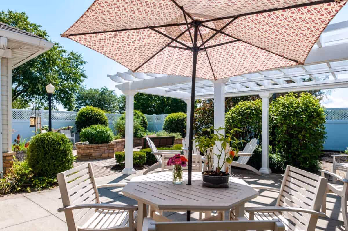 Shaded outdoor patio with a round table, umbrella and chairs under a pergola amid landscaped bushes.