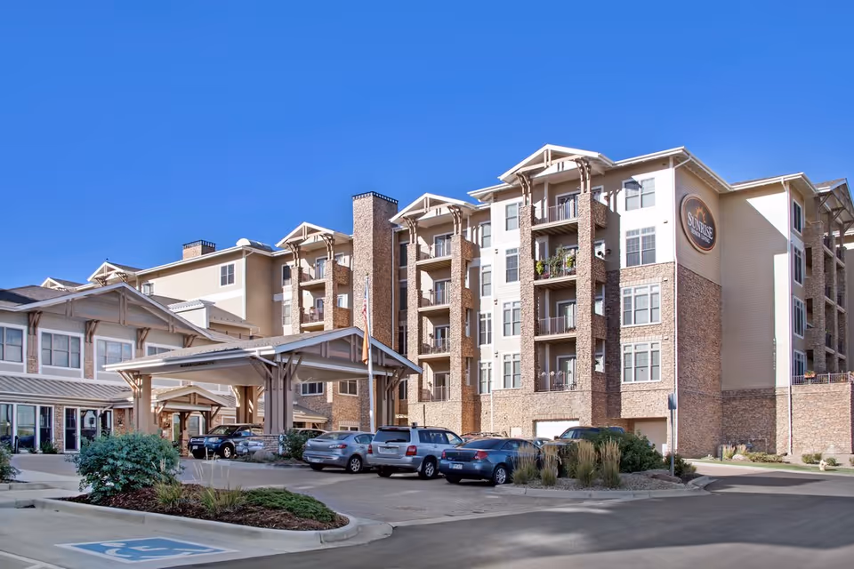 Exterior front view of the Sunrise at FlatIrons multi-story senior living building with a covered porte-cochere, parked cars, and a clear blue sky.