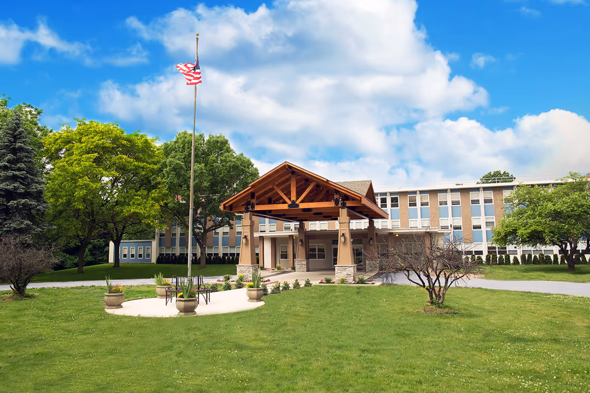 Front exterior view of Rochester Center, a multi-story building with a covered entrance supported by columns. There is a flagpole with an American flag in front, surrounded by a circular paved area with benches and planters. The building is surrounded by green grass, trees, and a partly cloudy blue sky.