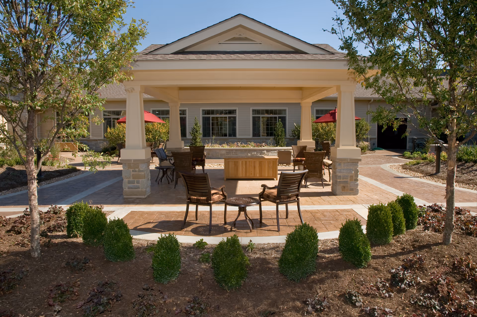 Covered outdoor patio with chairs, tables, umbrellas and landscaping in front of a senior living building.