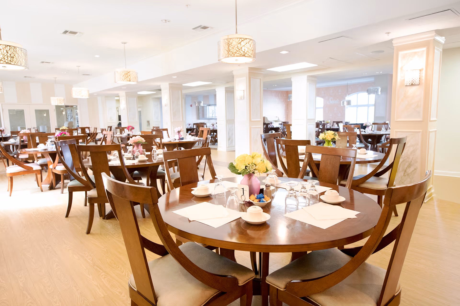 A bright and spacious dining room with multiple round wooden tables and matching chairs. Each table is set with white cups, saucers, glasses, napkins, and small flower arrangements in vases. The room features light-colored walls, wooden flooring, and modern hanging light fixtures.