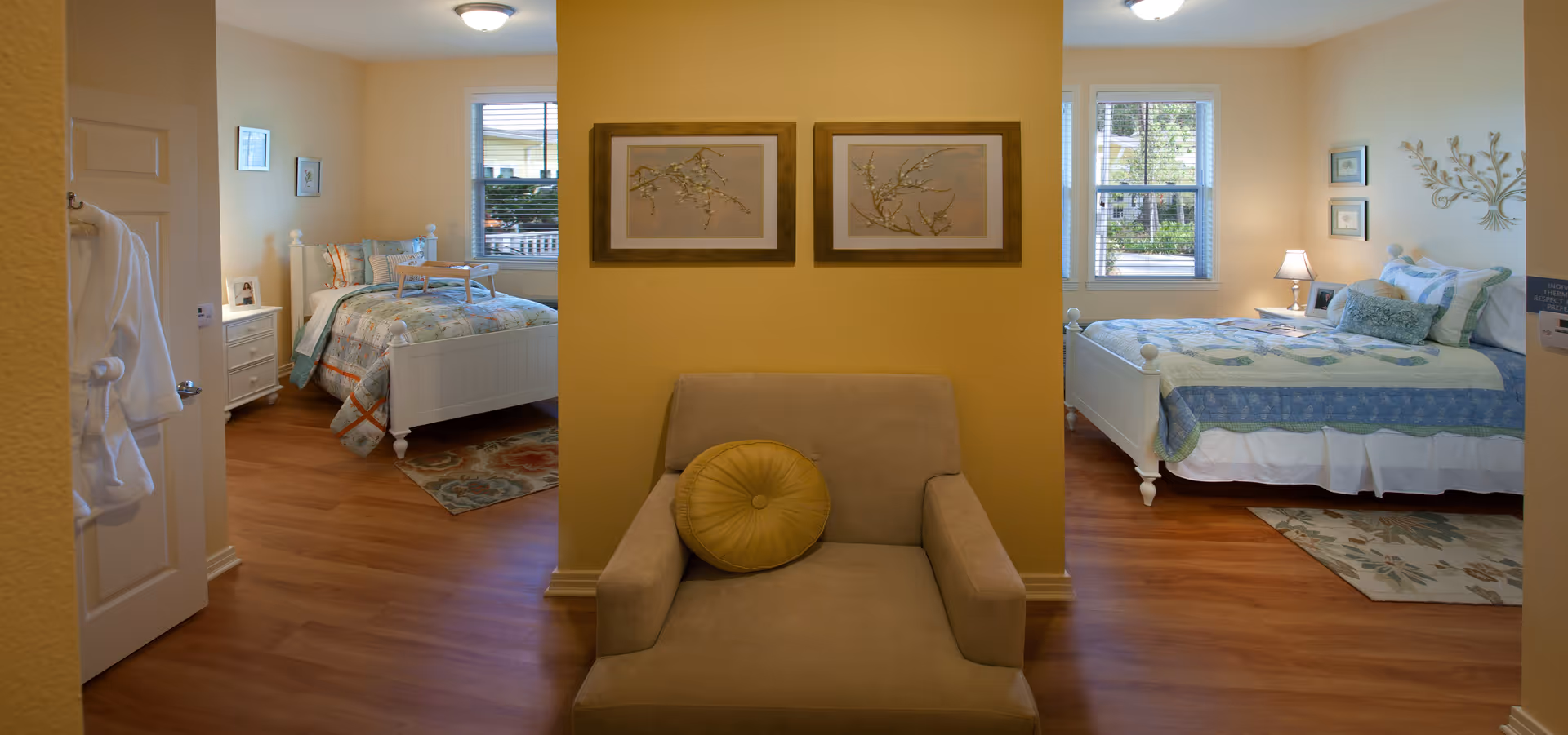 Interior view of a senior living facility room at Serenades at Longwood showing two separate bedrooms divided by a partial wall. Each bedroom has a bed with patterned bedding, a window with blinds, and bedside tables with lamps. In the center, against the dividing wall, there is a beige armchair with a round yellow cushion. The walls are painted light yellow and the floor is wooden.