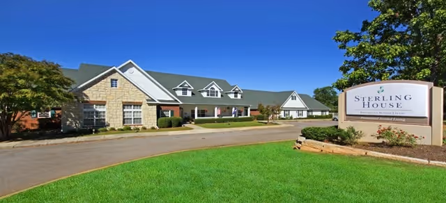 Front exterior of a senior living building with a driveway, manicured lawn, and a sign reading "Sterling House" under a clear blue sky.