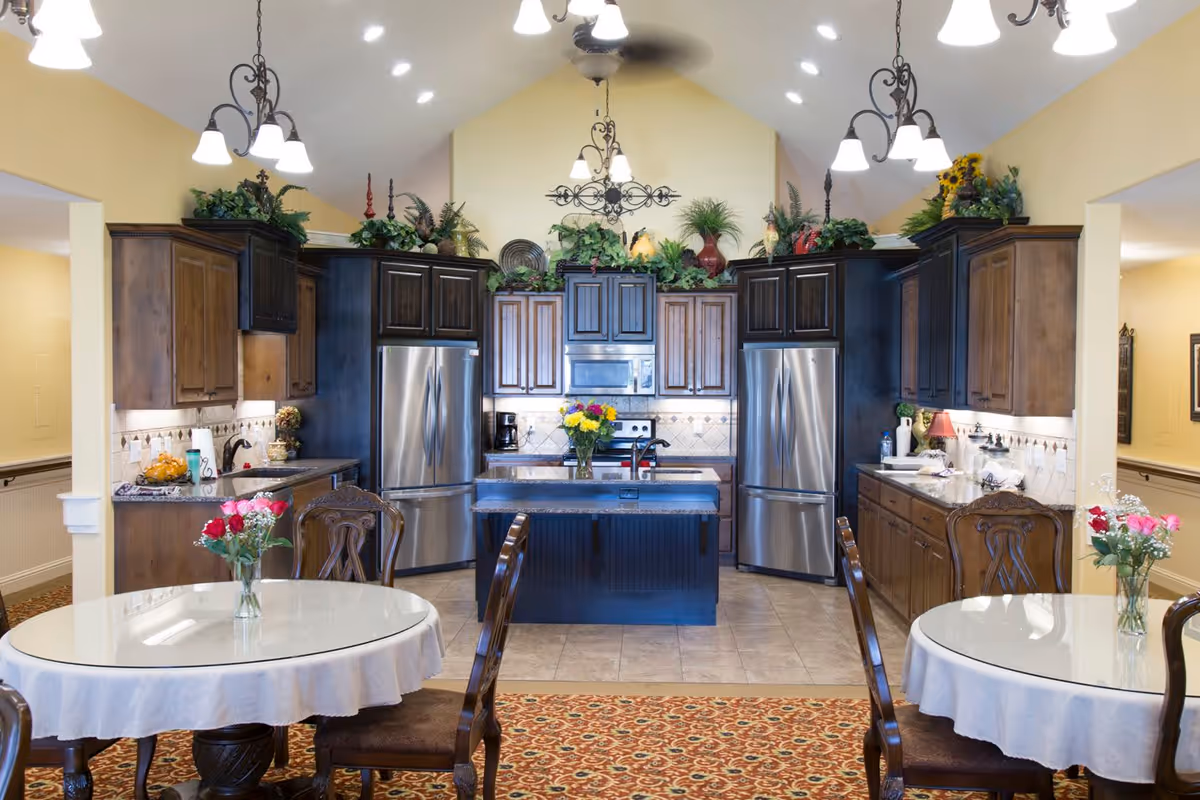 A spacious kitchen area with dark wood cabinets, two stainless steel refrigerators, a central island with a vase of flowers, and two round dining tables with white tablecloths and floral centerpieces. The ceiling is vaulted with multiple hanging light fixtures, and decorative greenery is placed above the cabinets.