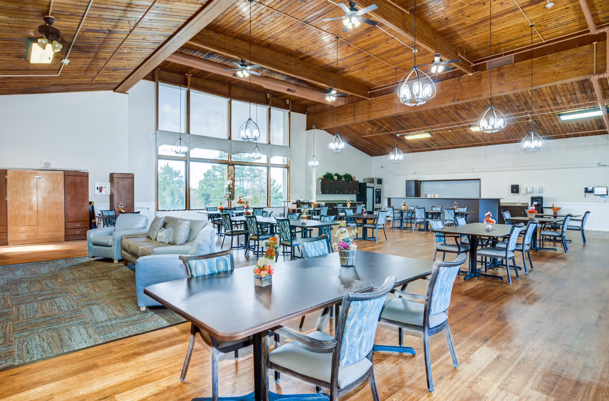 Spacious dining room with multiple tables and chairs arranged neatly. The room features a high wooden ceiling with ceiling fans and hanging light fixtures. Large windows allow natural light to fill the space. There is a seating area with a sofa and armchairs on the left side, and a counter area with cabinets and appliances in the back.