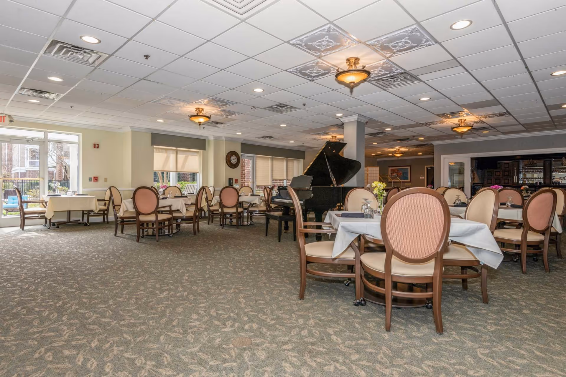 Spacious dining room with round tables and chairs, a grand piano, and large windows letting in natural light.
