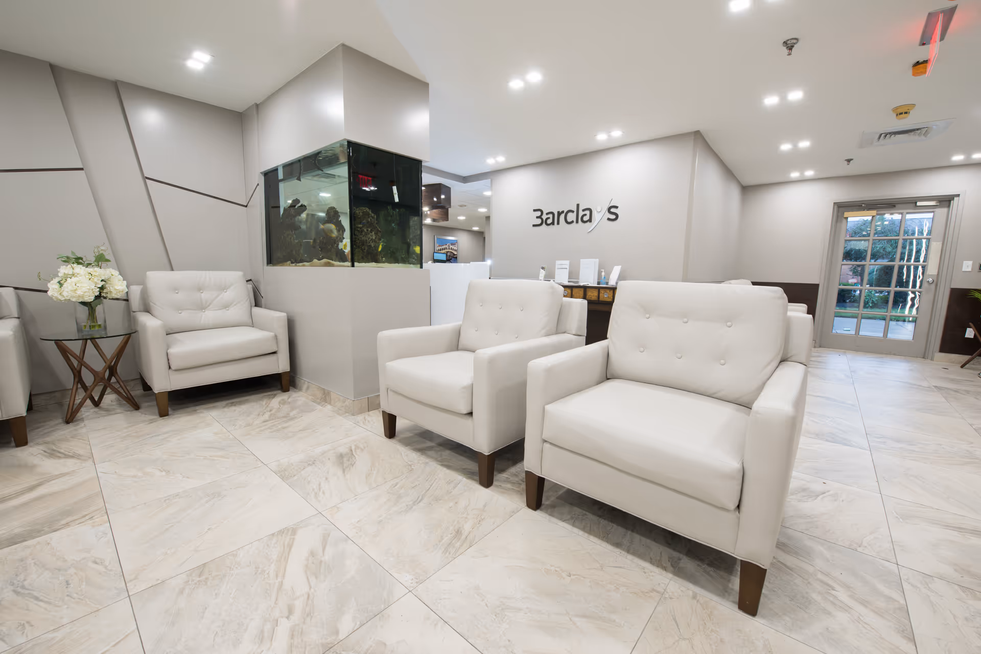 A modern and clean waiting area with beige armchairs arranged around a corner. There is a small glass-top side table with a vase of white flowers. A large built-in aquarium is embedded in the wall. The wall behind the reception desk displays the name 'Barclays'. The floor is tiled with light-colored marble, and a glass door leads outside.