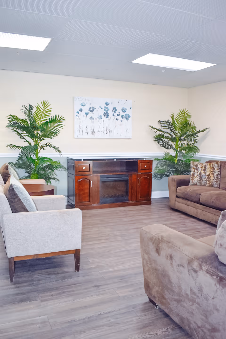 A cozy living room area with a wooden floor, featuring a brown sofa, a beige armchair, a wooden cabinet with an electric fireplace, two green potted plants, and a floral painting on the wall.