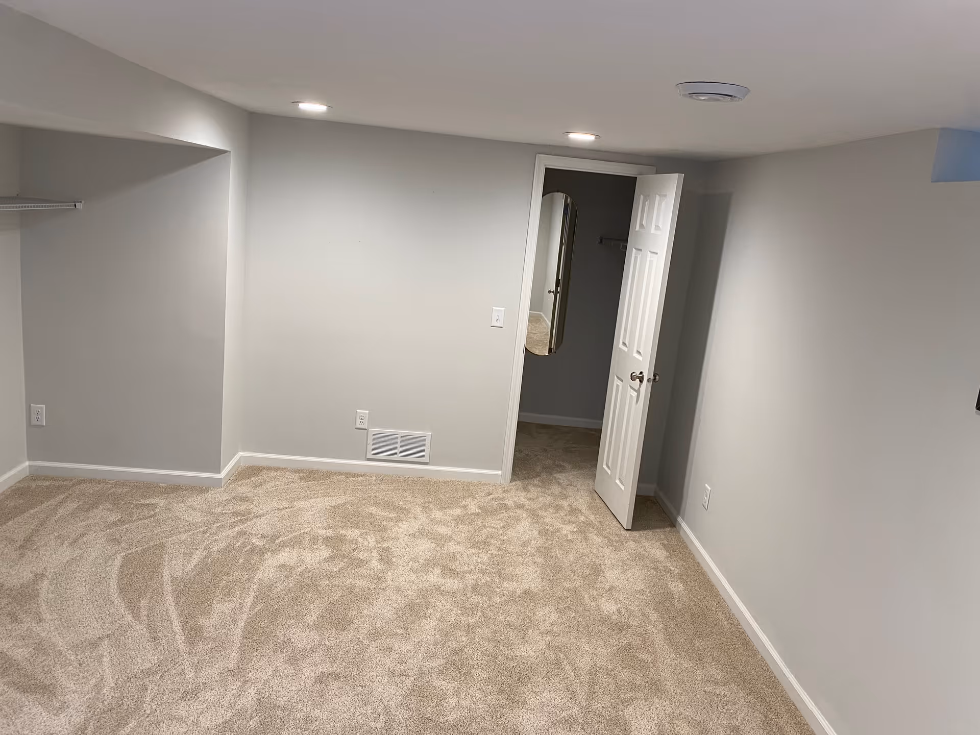 Empty carpeted room with light gray walls, recessed ceiling lights, and an open door revealing a small closet.