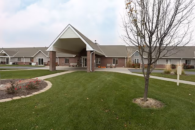 Exterior view of a single-story senior living facility building with a covered entrance supported by brick pillars, surrounded by a well-maintained lawn, a tree without leaves, and a small flower bed.