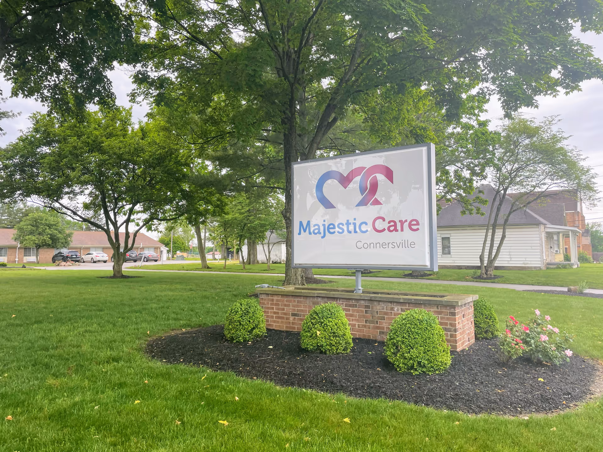 Majestic Care Connersville sign on a brick base in a landscaped lawn with trees and buildings behind it.