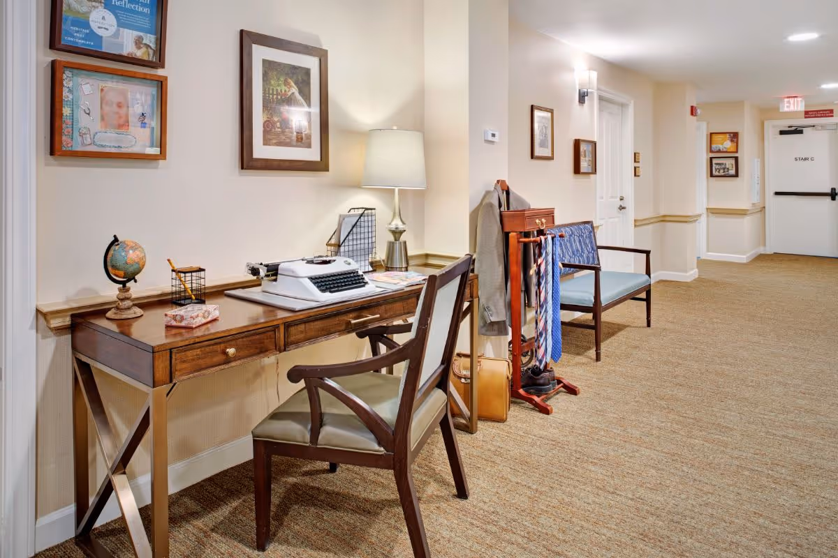 A hallway in a senior living facility featuring a wooden desk with a vintage typewriter, a globe, a lamp, and office supplies. Next to the desk is a wooden valet stand holding ties, a jacket, and shoes. A chair with blue upholstery is placed nearby. The hallway has beige walls, carpeted floors, framed pictures on the walls, and a door labeled 'STAIR C' at the end.