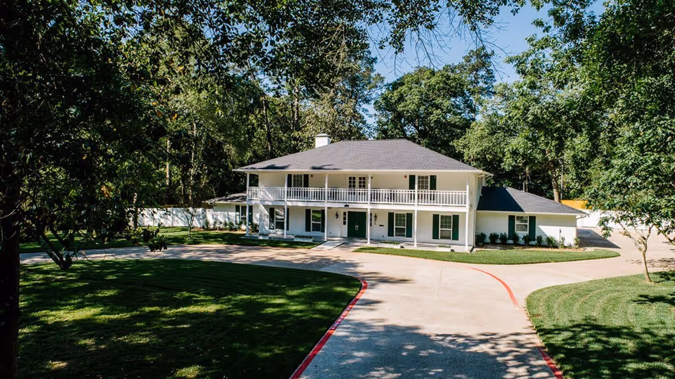 A large two-story white building with a dark gray roof surrounded by trees and greenery. The building has a balcony on the second floor and a driveway leading up to the front entrance. The area is well-maintained with trimmed grass and a curved concrete driveway bordered by a red line.