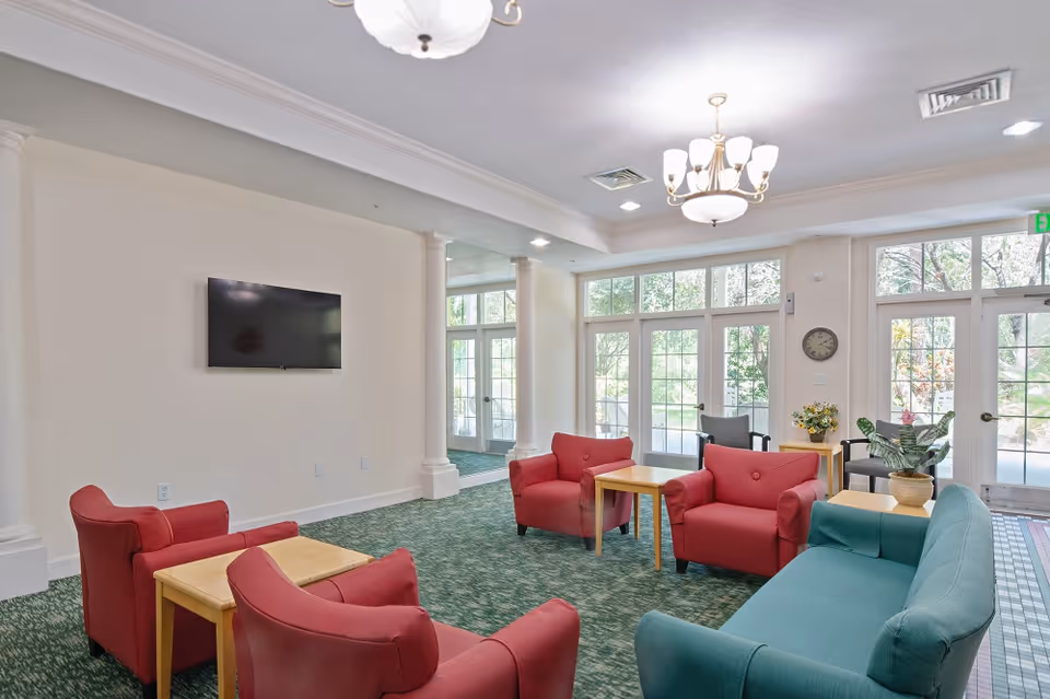 A bright and spacious living room area with large windows and glass doors letting in natural light. The room features red armchairs, a teal sofa, wooden side tables, a wall-mounted flat-screen TV, green carpet, and decorative plants. Two chandeliers hang from the ceiling.