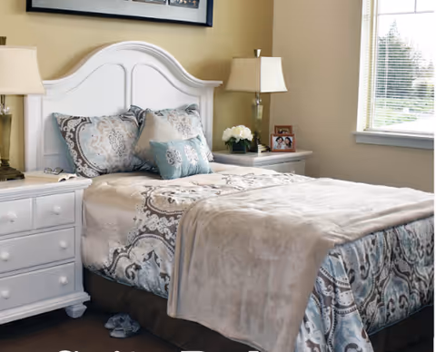 A neatly made bed with patterned bedding and multiple pillows in a bedroom. The bed has a white wooden headboard and is flanked by two white nightstands, each with a lamp. There is a window with blinds on the right side, and a framed photo and a vase with white flowers on the right nightstand.