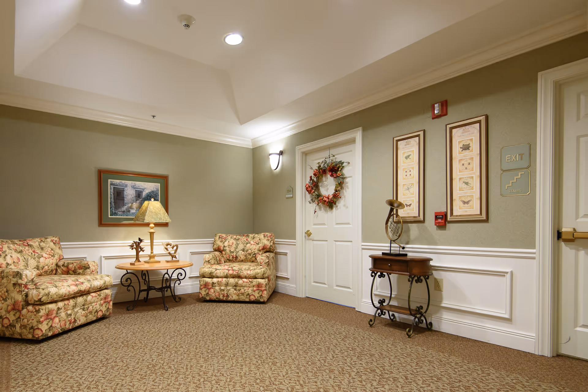 A cozy sitting area in a senior living facility hallway with two floral upholstered armchairs, a round wooden table with a lamp and decorative items, a white door adorned with a floral wreath, framed artwork on the walls, and a small decorative table with a brass horn sculpture.