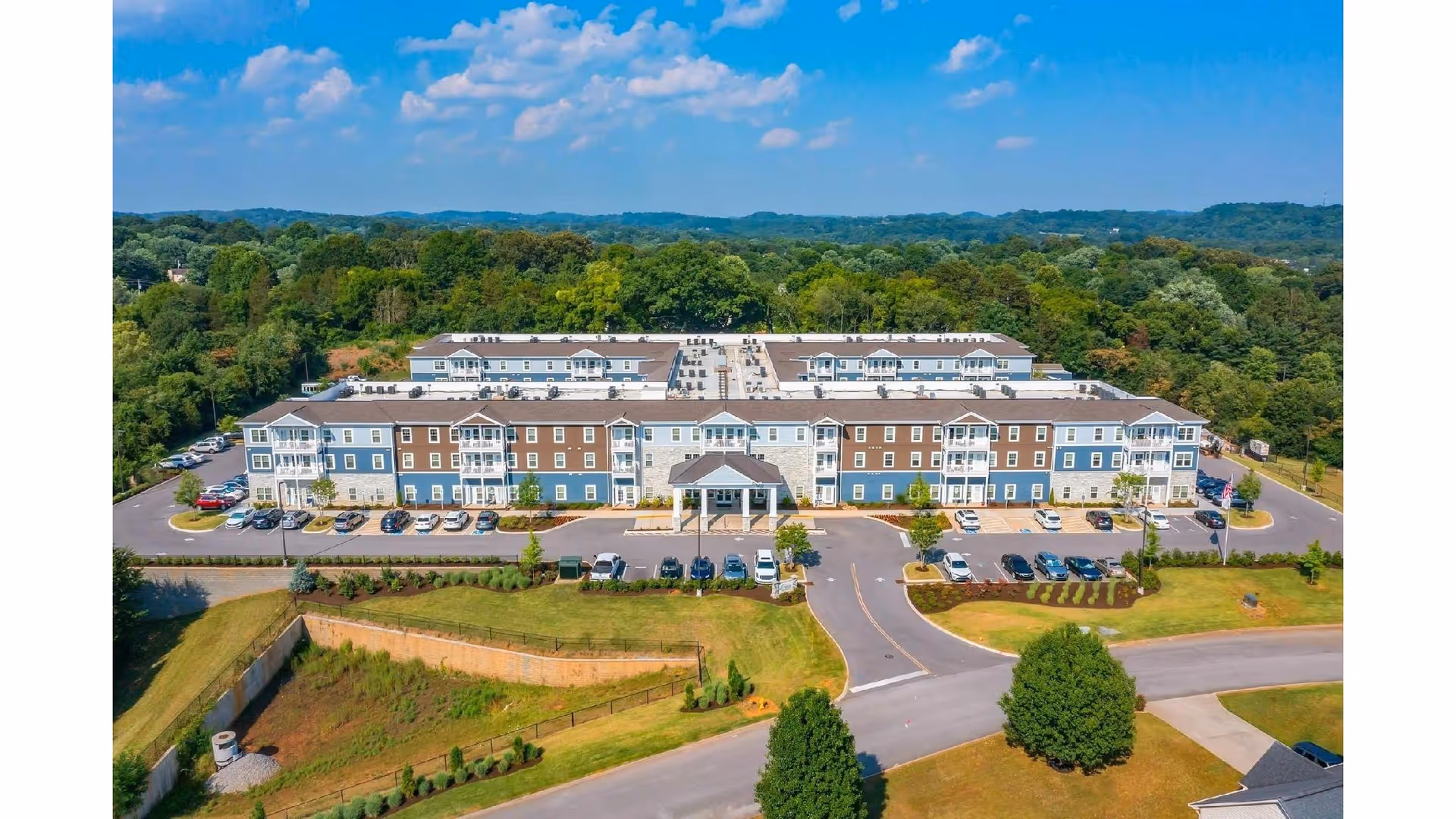 Aerial view of a large, multi-story senior living facility named Prime West Knoxville surrounded by greenery and trees under a blue sky with scattered clouds. The building has a mix of blue, brown, and white exterior walls with multiple balconies and a covered entrance. There are several cars parked in the parking lots in front of the building.