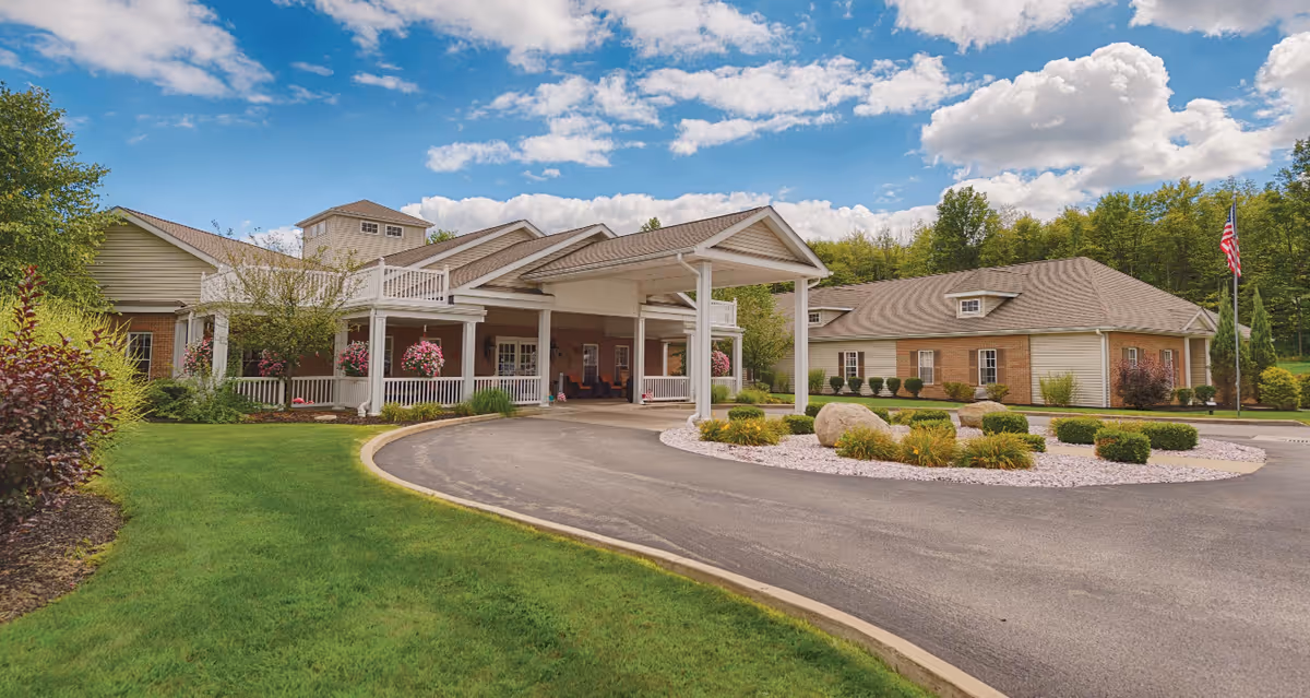 Exterior view of Victoria House Assisted Living facility showing a large building with a covered entrance, well-maintained landscaping including green grass, bushes, and flowers, a circular driveway, and an American flag on a flagpole under a partly cloudy blue sky.