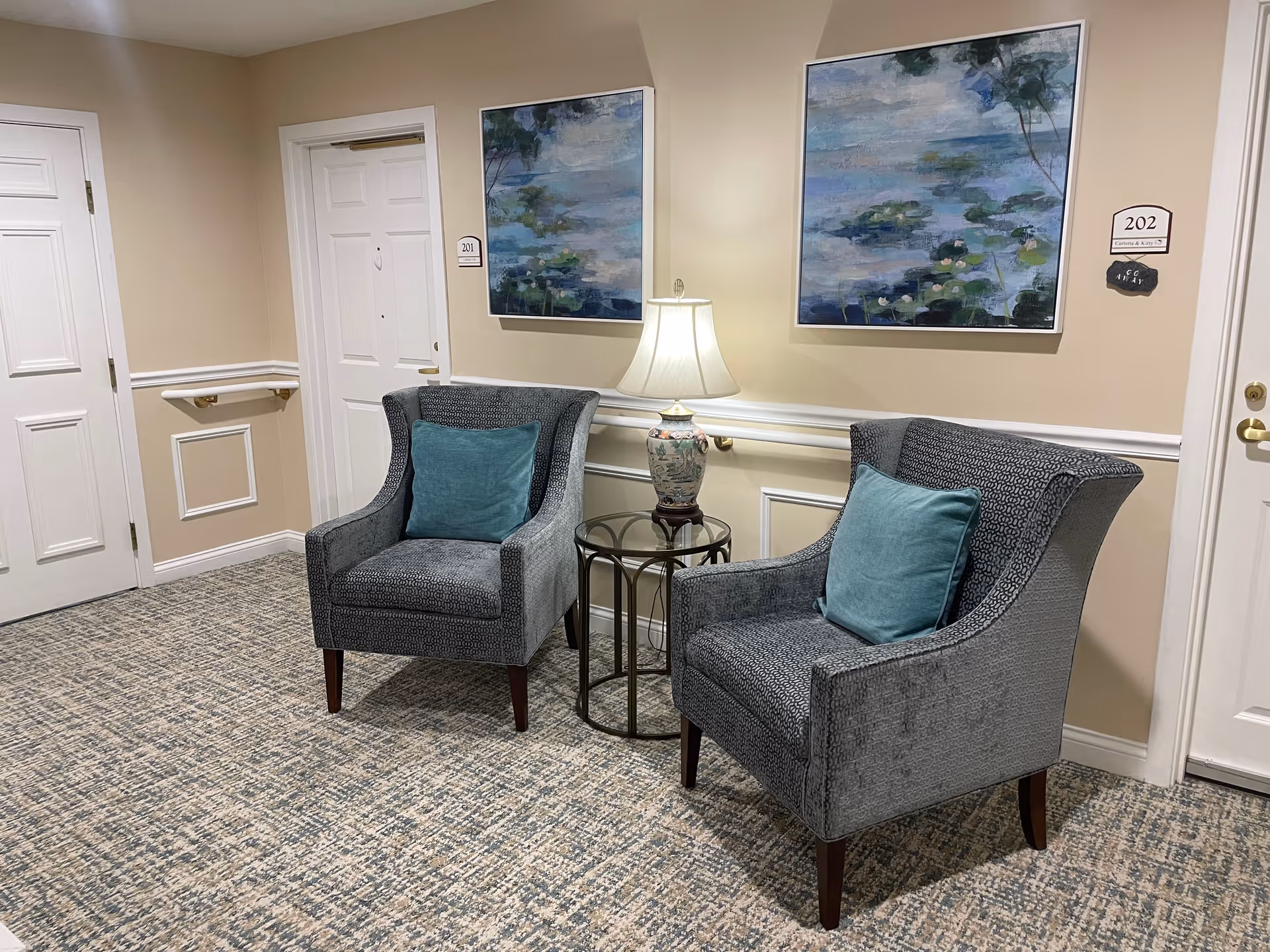 Two upholstered armchairs with teal pillows beside a small table and lamp in a hallway outside resident doors with framed artwork on the wall.