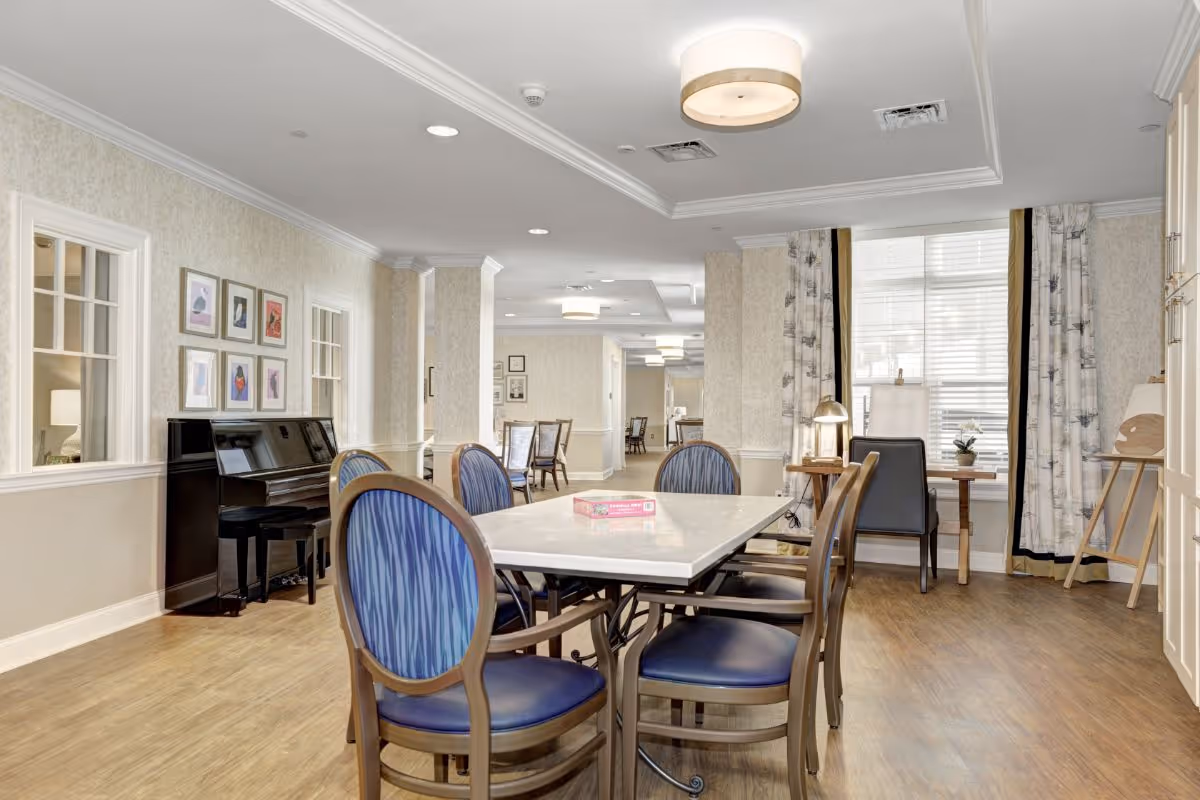 A bright and spacious common area in a senior living facility featuring a rectangular table with six blue cushioned chairs around it. In the background, there is a black upright piano against the wall with framed artwork above it. The room has light-colored walls, wood flooring, large windows with patterned curtains, and ceiling lights. A small desk with a chair and lamp is positioned near the window.