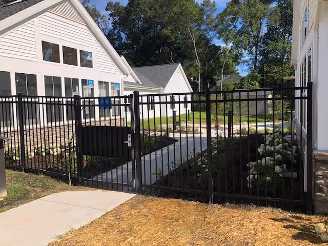 Black metal fence gate enclosing a landscaped pathway leading to white residential buildings with large windows, surrounded by green trees and shrubs under a clear sky.