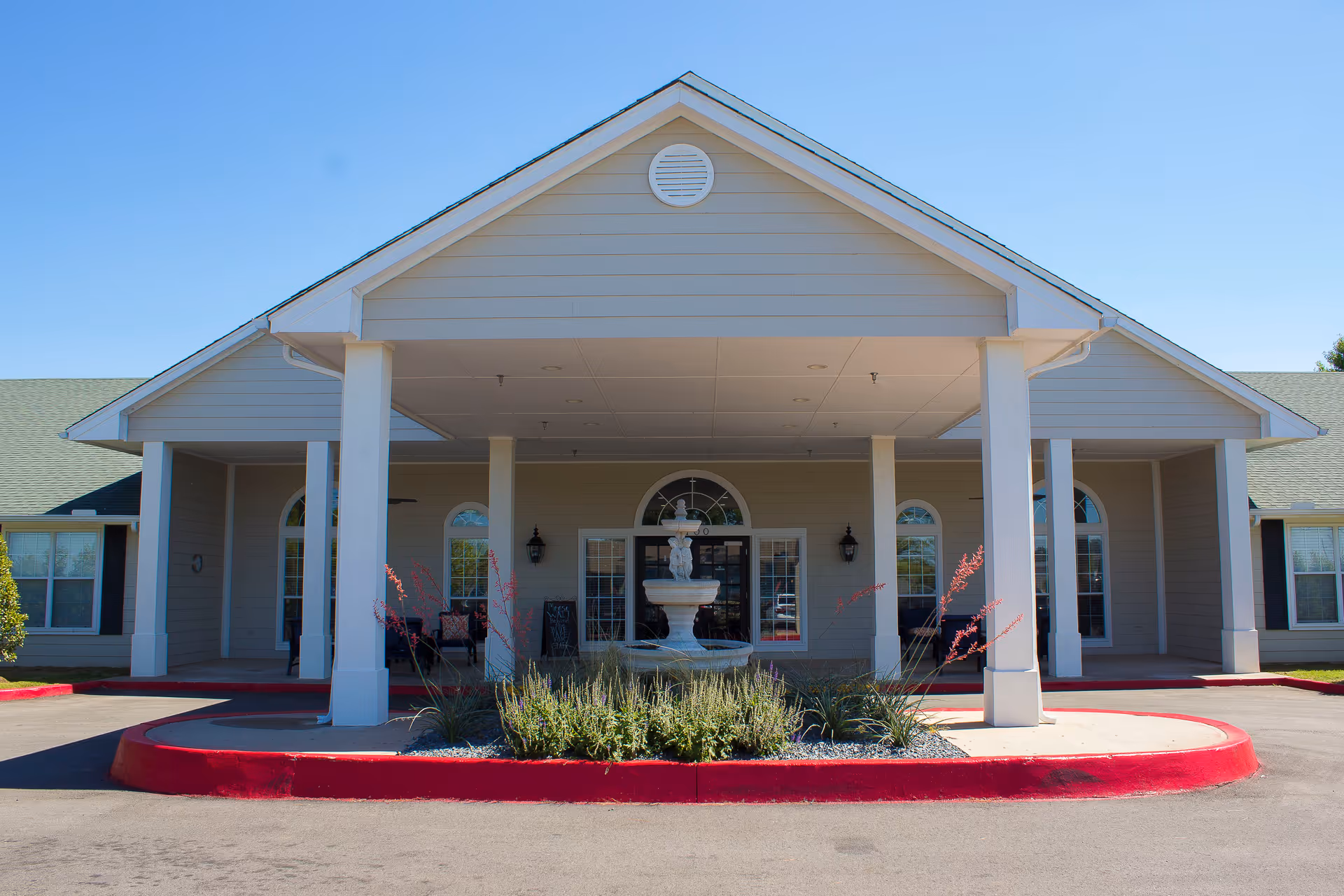Front exterior view of Hefner Mansions Senior Independent Living building with a covered entrance supported by white pillars, a central water fountain surrounded by plants, and clear blue sky in the background.