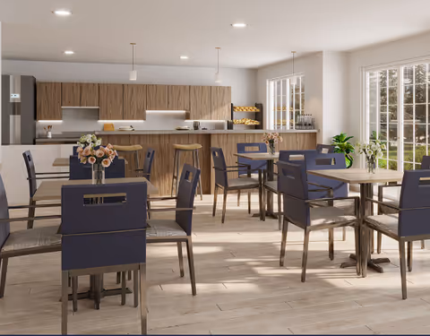 A bright and modern dining area with several tables and chairs arranged neatly. Each table has a small vase with flowers. In the background, there is a kitchen area with wooden cabinets, a refrigerator, and a counter with bar stools. Large windows allow natural light to fill the room, and a plant is placed near the window.