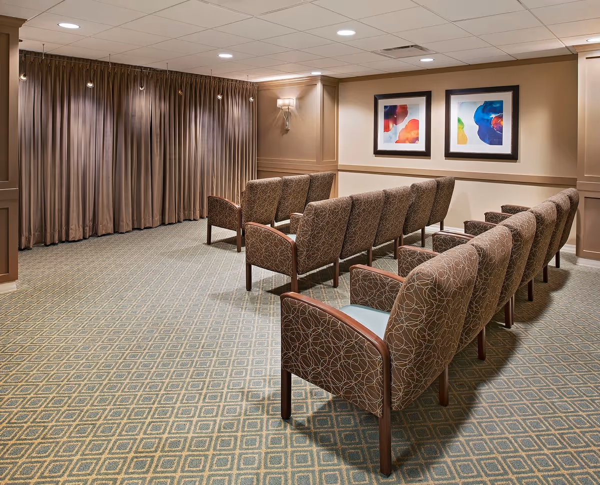 A small theater or presentation room with two rows of patterned upholstered chairs facing a wall with closed brown curtains. The room has beige walls with two colorful abstract paintings and soft lighting from ceiling lights and a wall sconce.