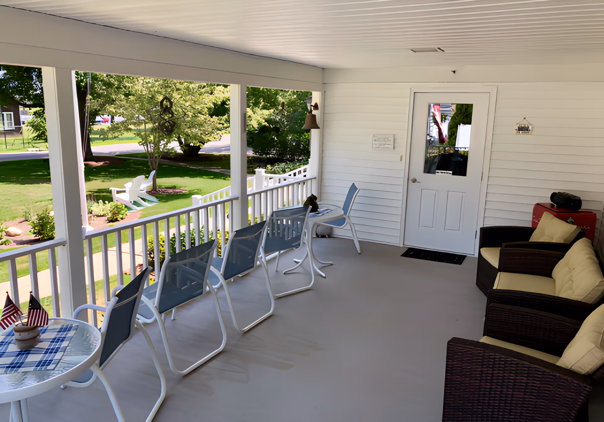 Covered porch with a row of chairs, small tables and wicker seating overlooking a grassy yard.
