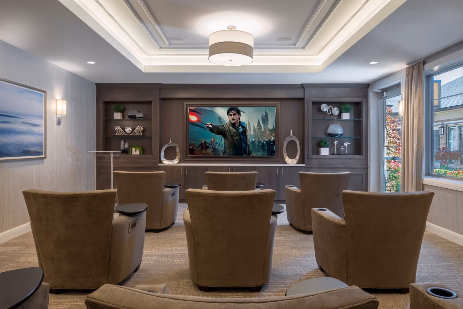 A cozy media room with six brown recliner chairs arranged in two rows facing a large flat-screen TV mounted on a dark wood built-in cabinet. The TV displays a scene from a Harry Potter movie. The room has a modern ceiling light fixture, built-in shelves with decorative items, a large window with beige curtains letting in natural light, and a framed ocean-themed artwork on the left wall.
