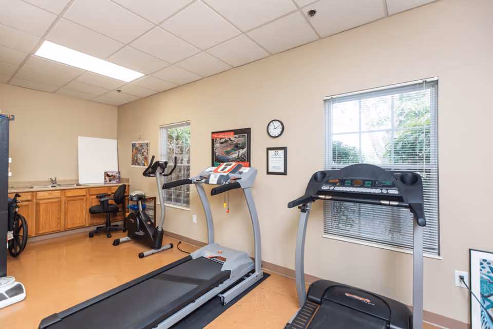 A small fitness room with two treadmills, an exercise bike, a wheelchair, and a chair near a countertop with a sink. The room has beige walls, two windows with blinds, a clock, and framed pictures on the wall.