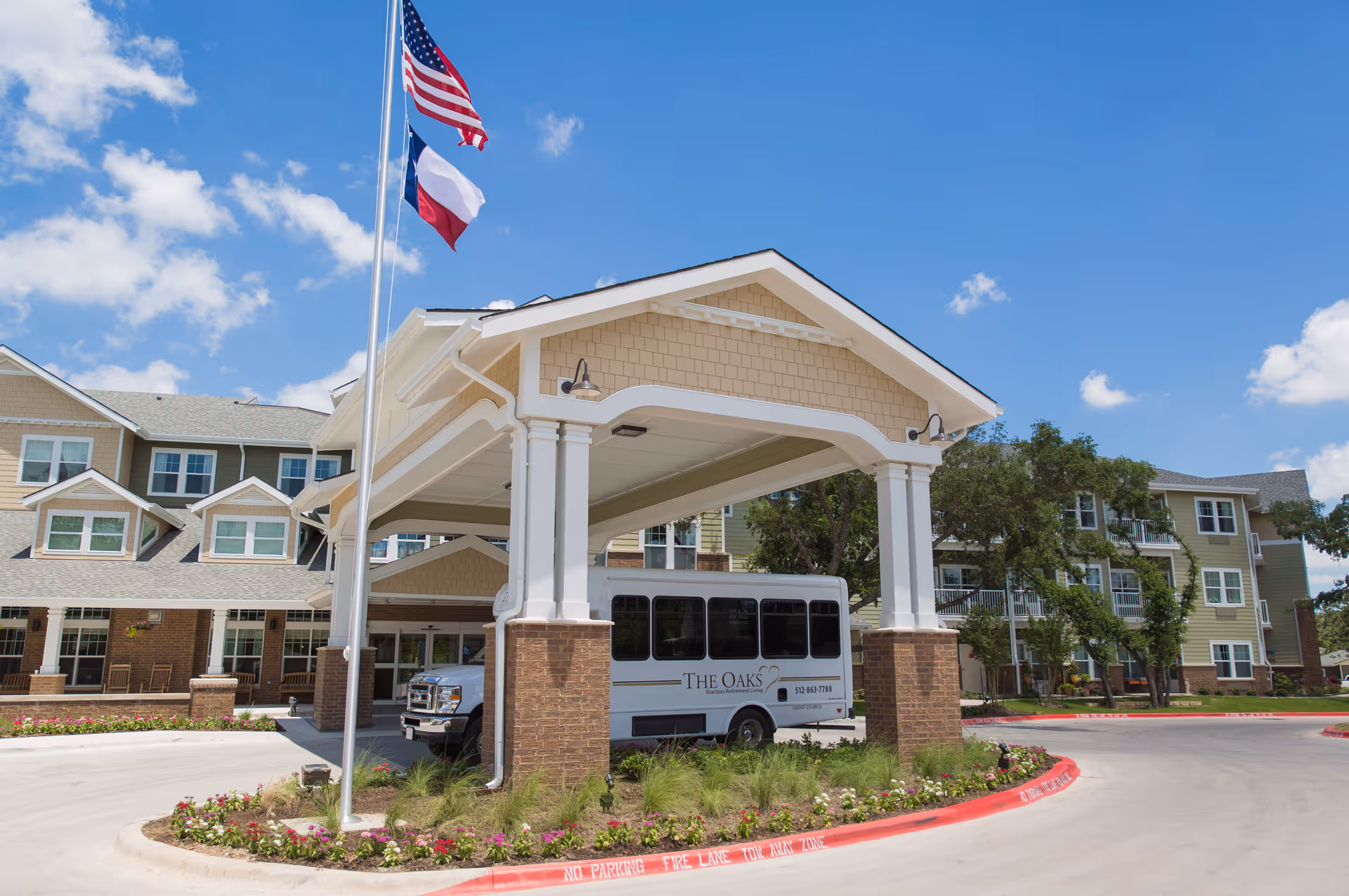 Exterior view of The Oaks Gracious Retirement Living facility showing the main entrance with a covered drop-off area, a shuttle bus parked underneath, and two flagpoles flying the American and Texas flags. The building is multi-story with beige siding and brick accents, surrounded by landscaping and a clear blue sky.