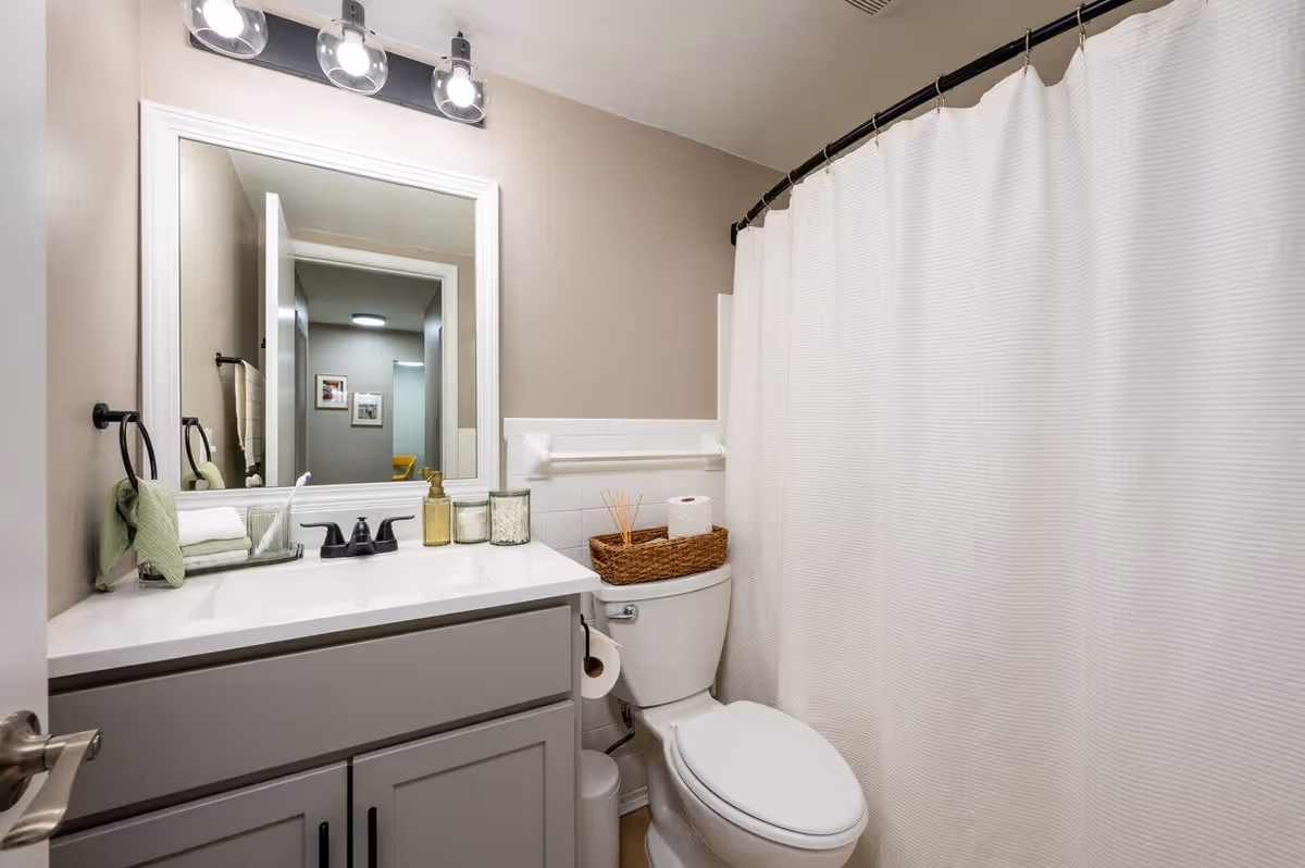 A modern bathroom with a sink vanity and mirror, a toilet topped with a wicker basket, and a shower with a white curtain.