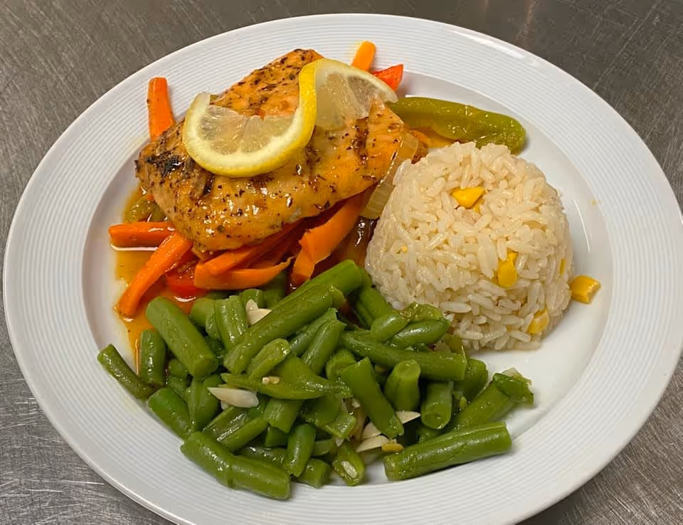 A white plate with a serving of cooked green beans with sliced almonds, a portion of white rice mixed with corn kernels, and a piece of grilled fish topped with two lemon slices, served on a bed of cooked mixed vegetables including carrots and bell peppers.