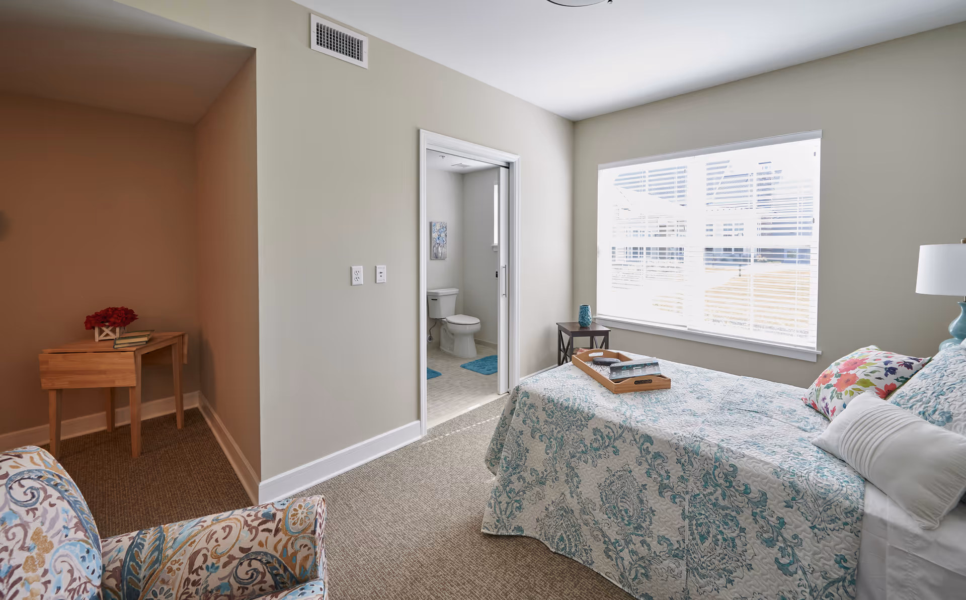 Sunlit bedroom with a patterned bedspread, armchair, small side table and an open door revealing an ensuite bathroom.