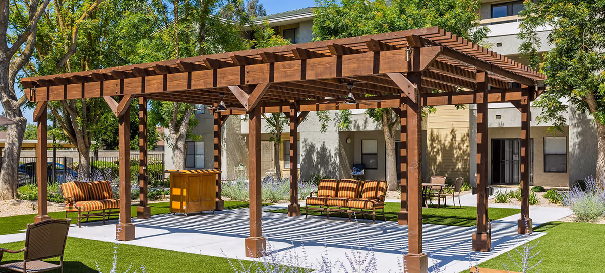 Wooden pergola shading striped cushioned benches and chairs on a paved patio in a landscaped courtyard of an apartment-style senior living building.