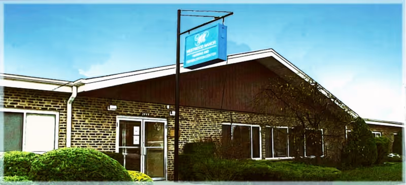 Exterior view of a single-story brick building with a peaked roof and several windows. There is a sign hanging from a pole near the entrance that reads Westwood Manor Nursing and Rehabilitation Center. Bushes and greenery are visible in front of the building under a clear blue sky.